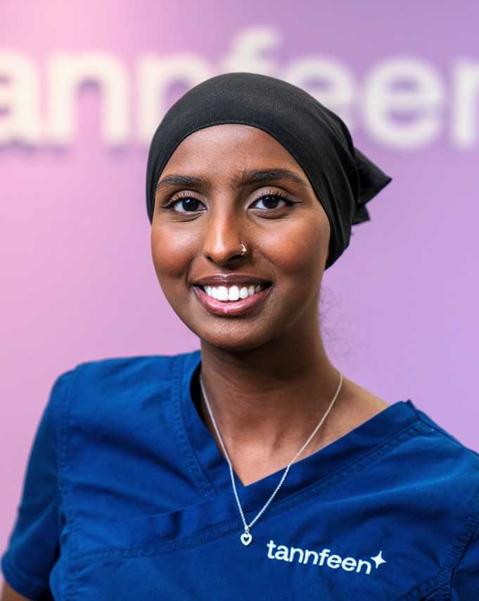 Portrett av en smilende kvinne i blå tannlegeuniform med «tannfeen» logo, svart hijab, nesering og et hjerteformet halskjede, mot en lilla gradientbakgrunn.