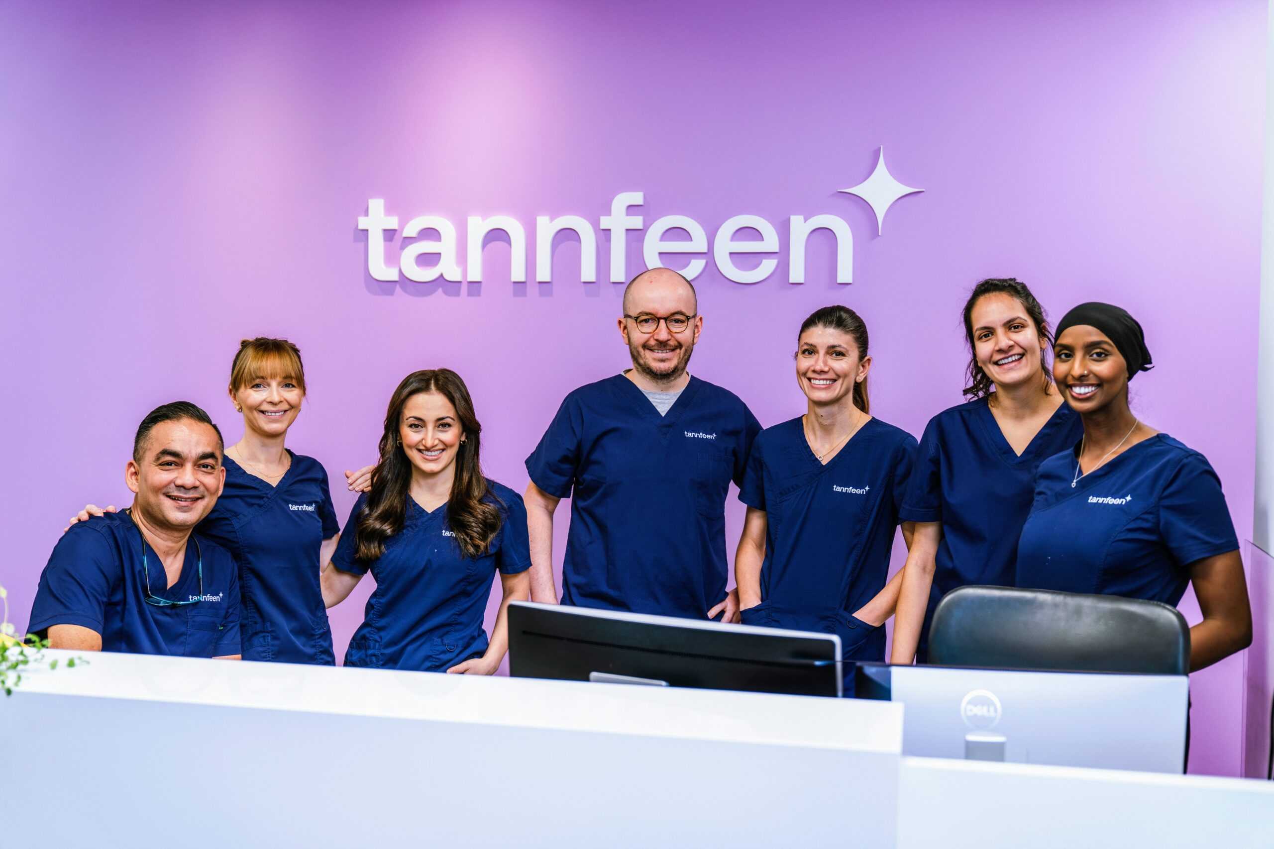 Syv tannlege-/klinikkmedarbeidere i mørkeblå uniform står samlet bak en resepsjon, foran en lilla vegg med logoen «tannfeen» og en stjerne, alle smiler mot kamera.