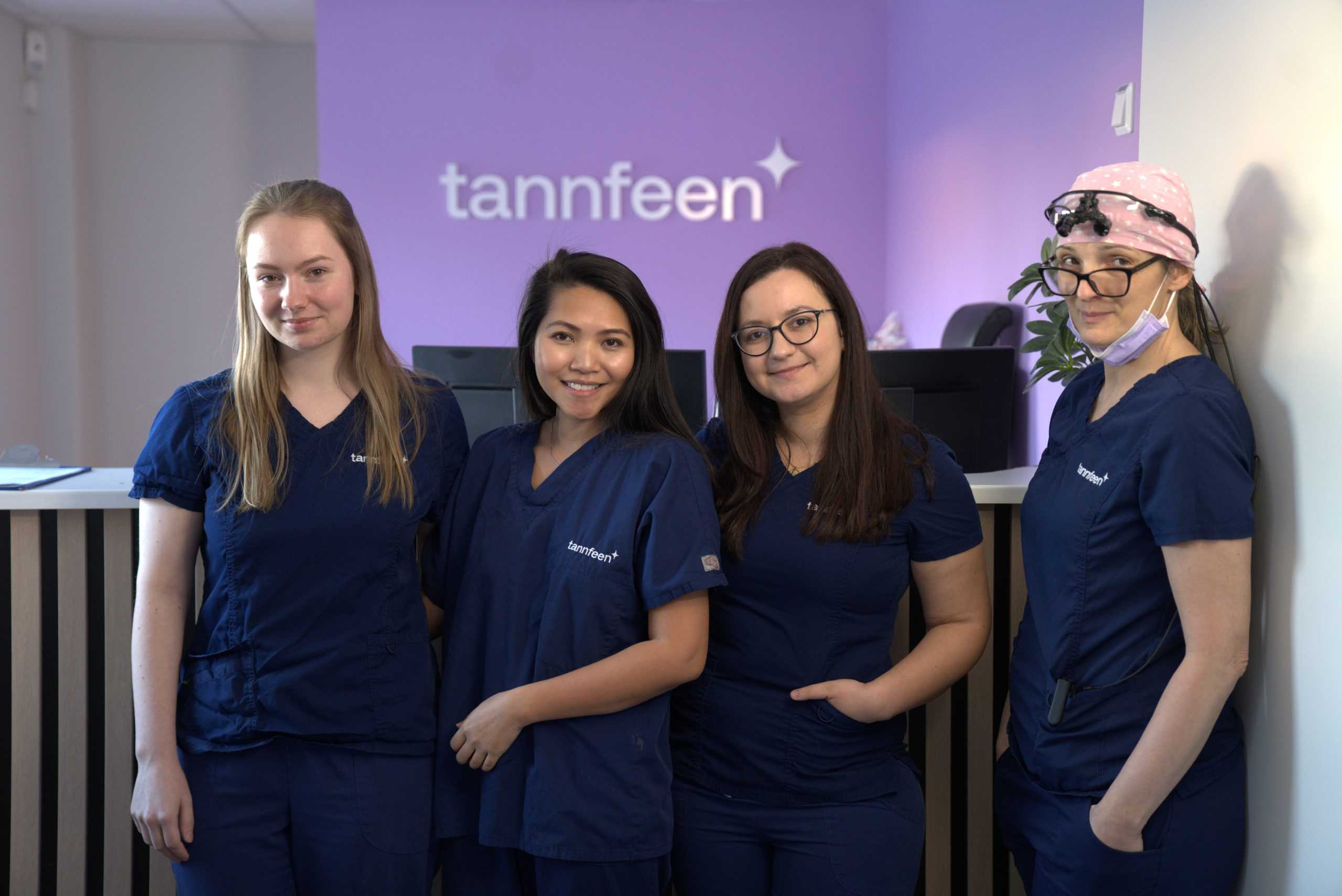 Fire kvinner i blå tannlegeuniformer står side om side ved resepsjonen i en tannklinik, foran en lilla vegg med logoen «tannfeen».