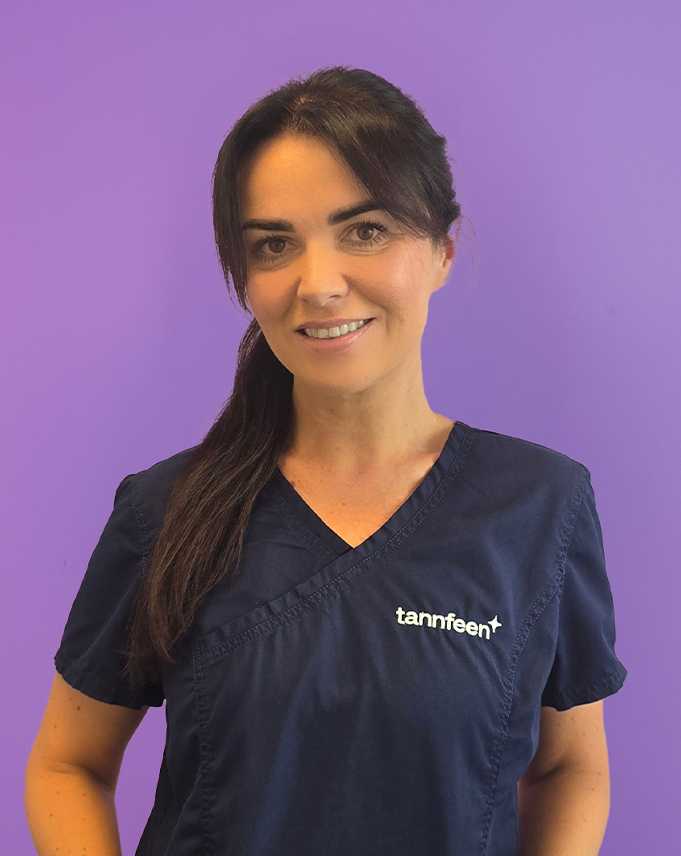 Kvinne i en mørkeblå tannlegeuniform står foran en lilla bakgrunn og smiler; på brystet står den broderte logoen «tannfeen».