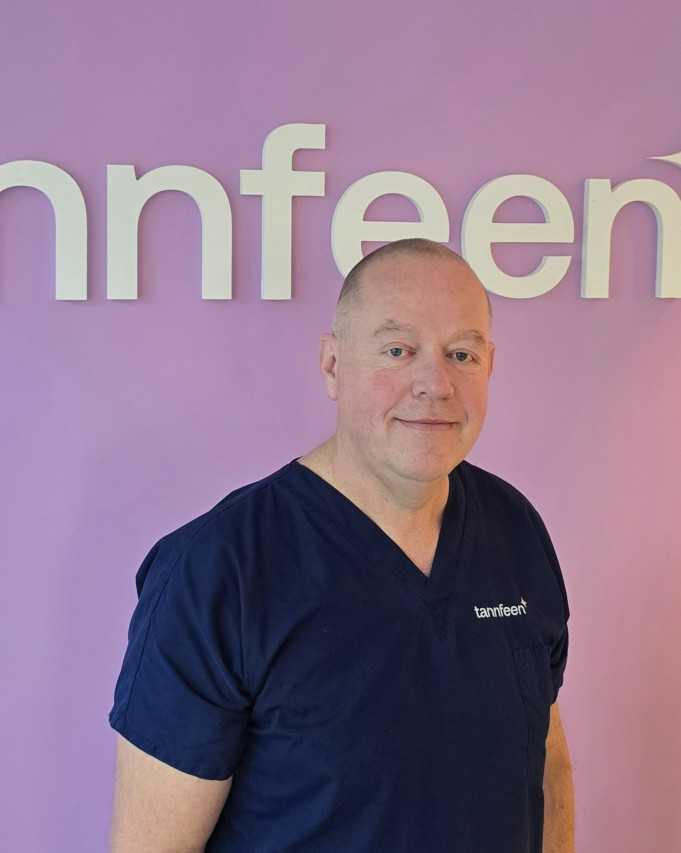 Portrett av en middelaldrende mann i mørkeblå medisinsk uniform (scrubs) som står foran en rosa-lilla vegg med store hvite bokstaver; han smiler litt mot kamera.