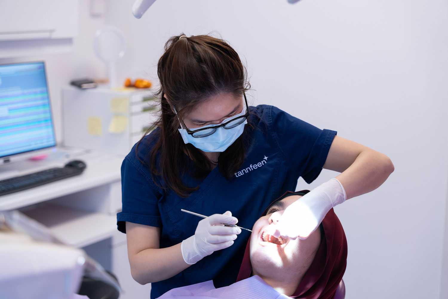 En tannlege eller tannpleier i blå uniform, ansiktsmaske og hansker utfører en tannbehandling på en pasient som ligger i tannstolen. Pasienten har munnåpning og burgunderfarget hodeplagg; i bakgrunnen ses dataskjerm og kontorutstyr.
