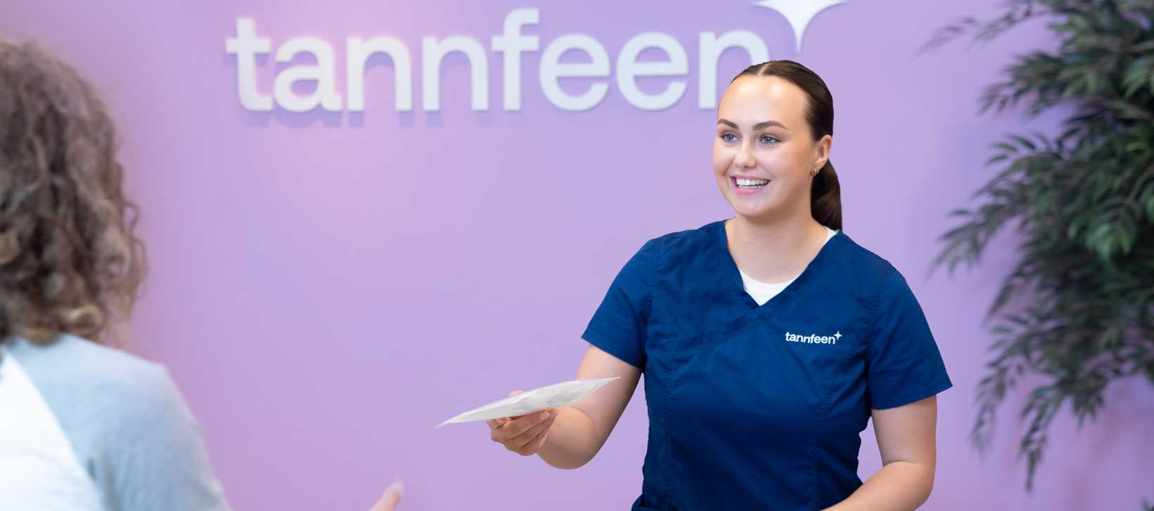 Kvinne i blå tannlegeuniform smiler mens hun gir et ark til en pasient. Bakgrunnen er en lilla vegg med logoen “tannfeen” og grønne planter.