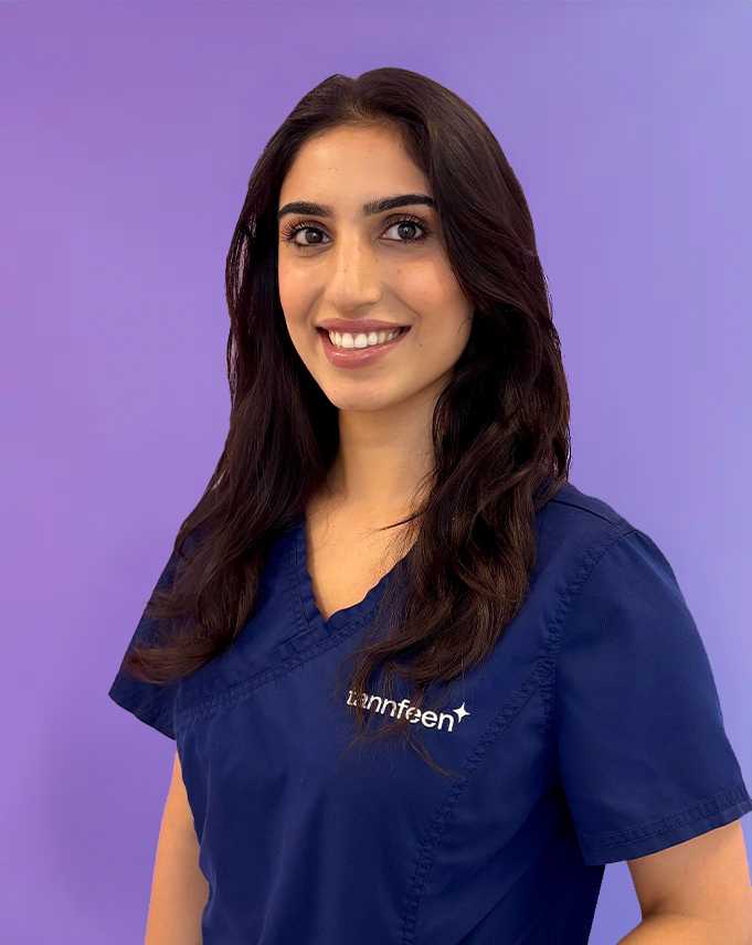 Portrett av en kvinne med mørk, bølget hår i blå sykepleieruniform, smilende mot en lilla bakgrunn, med hvitt logo på venstre bryst.